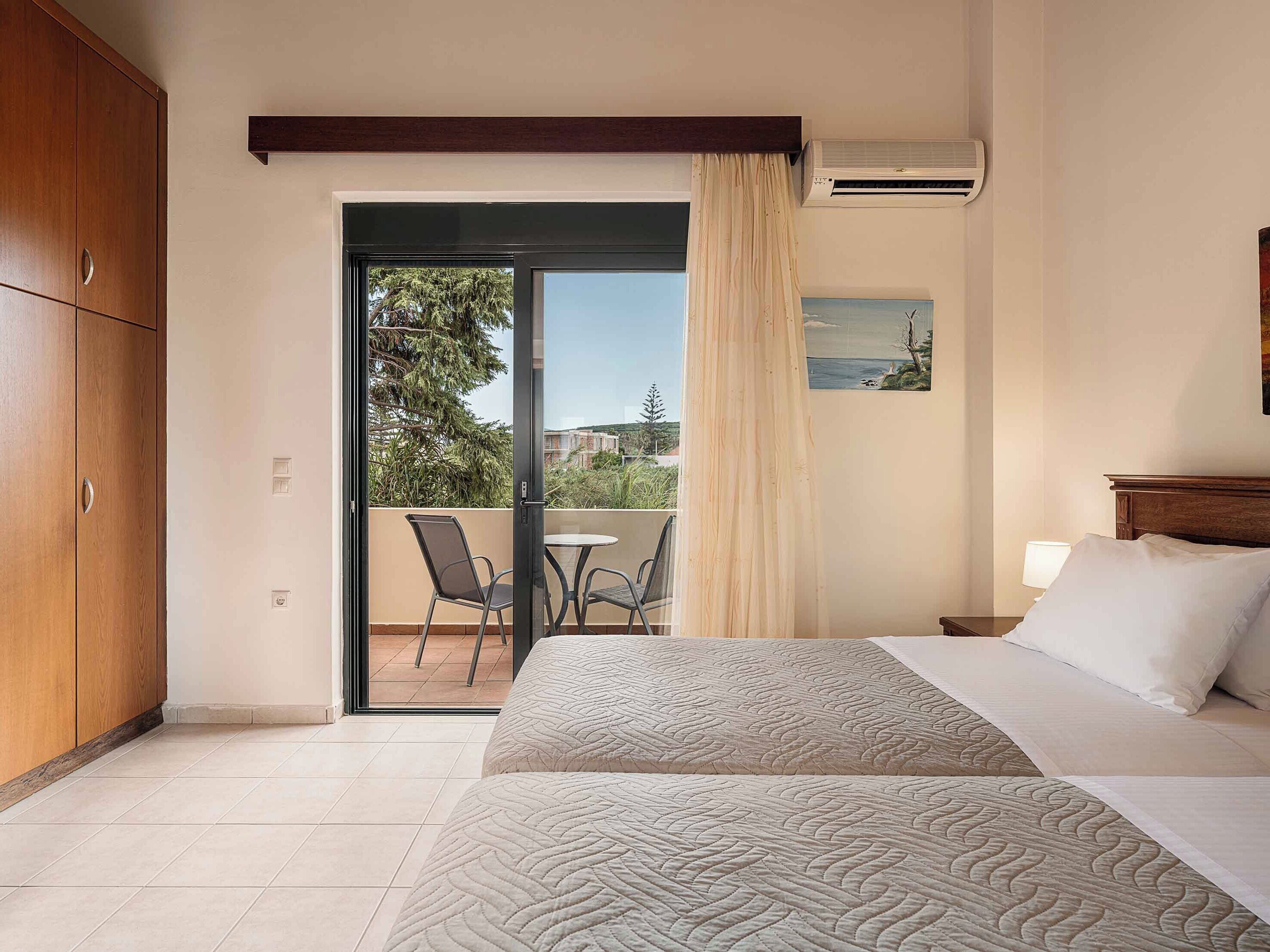 A hotel room with a bed, a balcony with chairs, a wooden wardrobe, and an air conditioning unit.