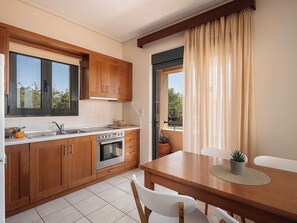 A modern kitchen with wooden cabinets, a built-in oven, and a dining area with a table and chairs. - Corali Villas West-Seaview, 2 Pools, 450m to Beach (Chania)