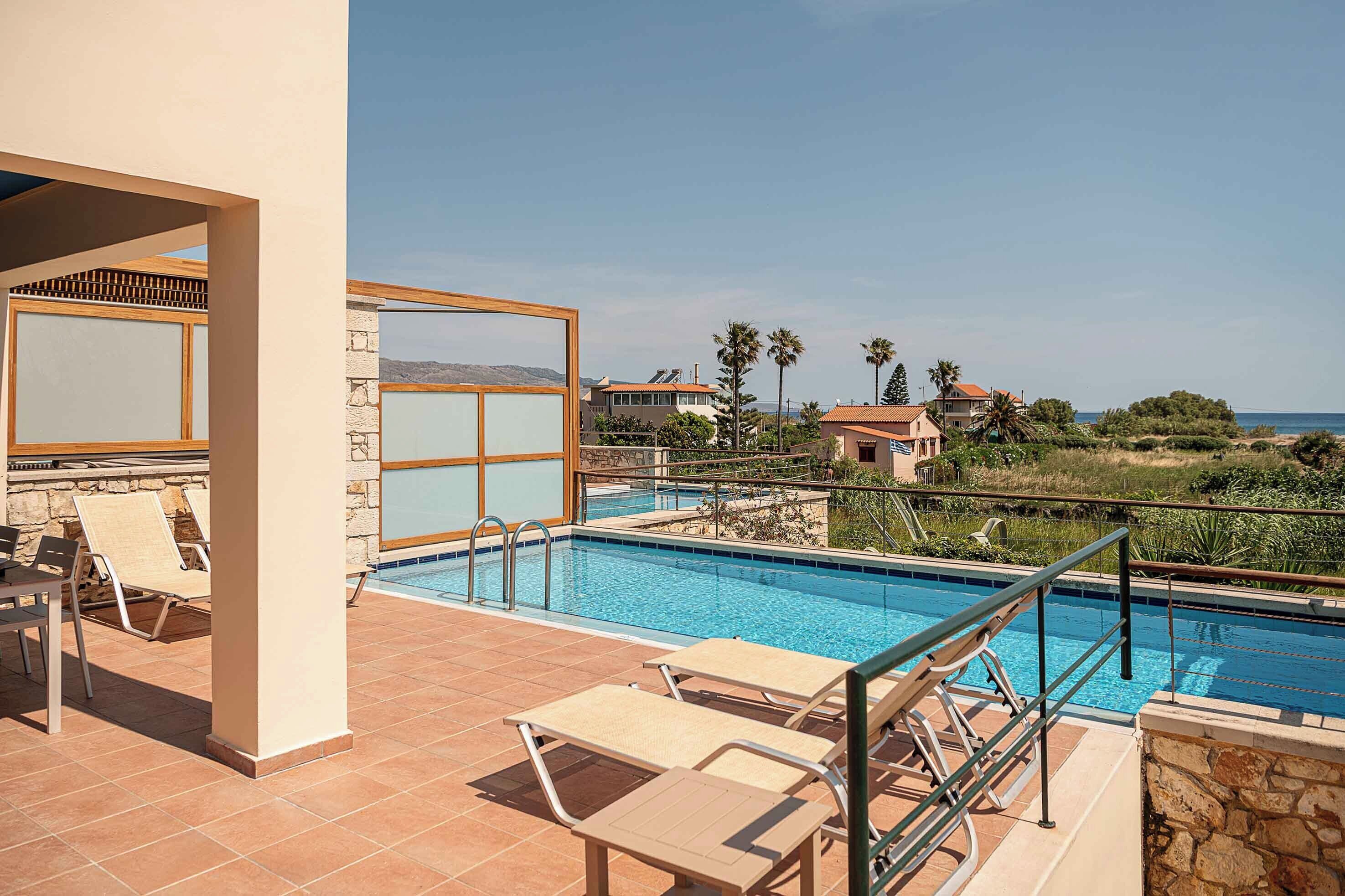 Una piscina con sillones de descanso y un muro de piedra, con vista a una zona residencial con casas y palmeras.