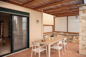 An outdoor patio with a wooden table and chairs, a stone wall, and a sliding glass door leading to an interior space. - Corali Villas Complex-Seaview,4 Pools,450 to Beach (Chania)