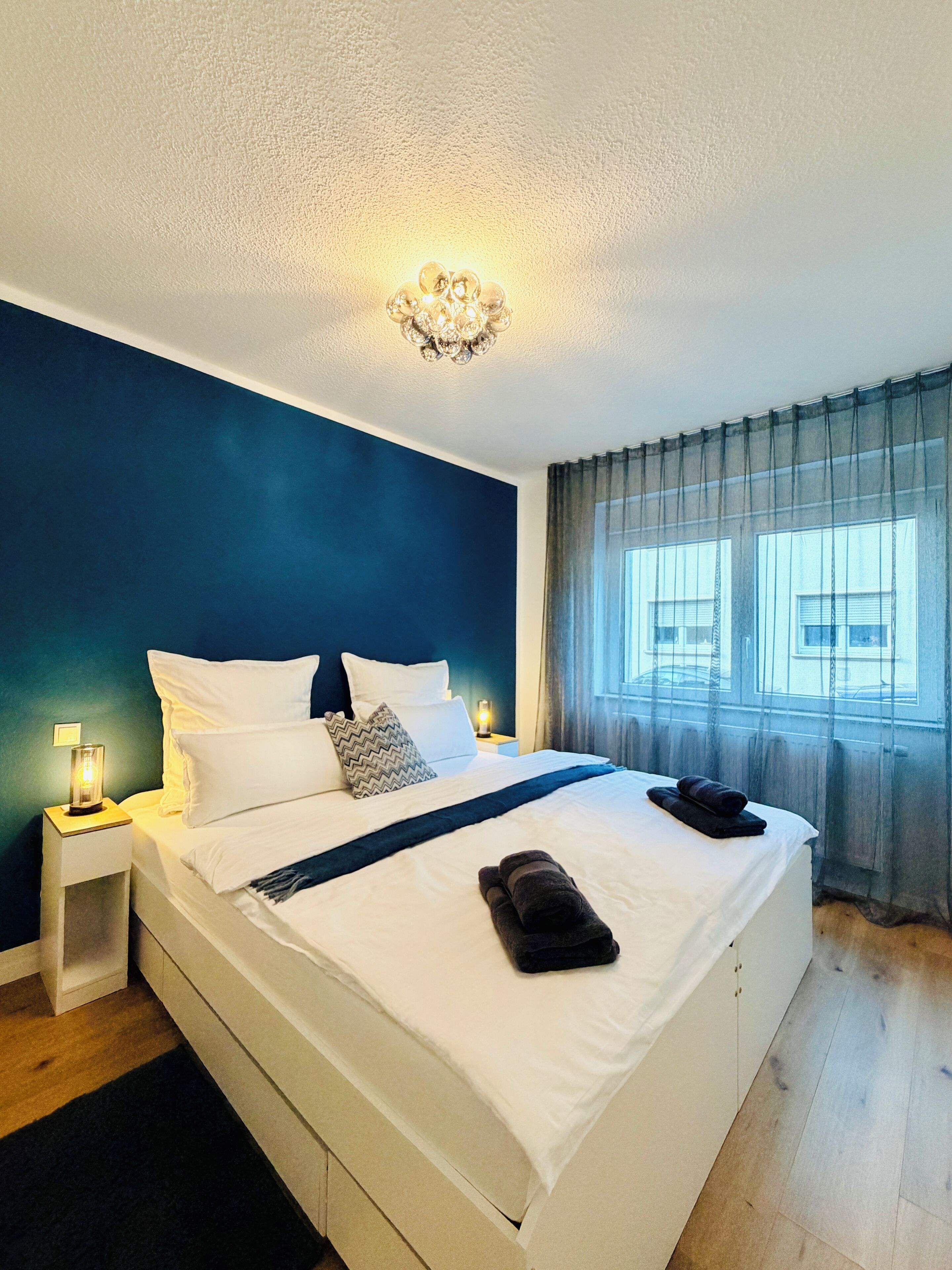 A bedroom with a large bed, white bedding, a nightstand with a lamp, and a view of the outside through sheer curtains.