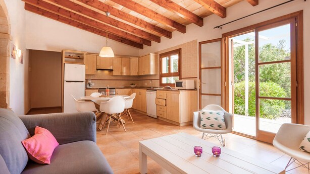 A modern kitchen with wooden beams, a dining area with a white table and chairs, and a large window with a view of greenery.