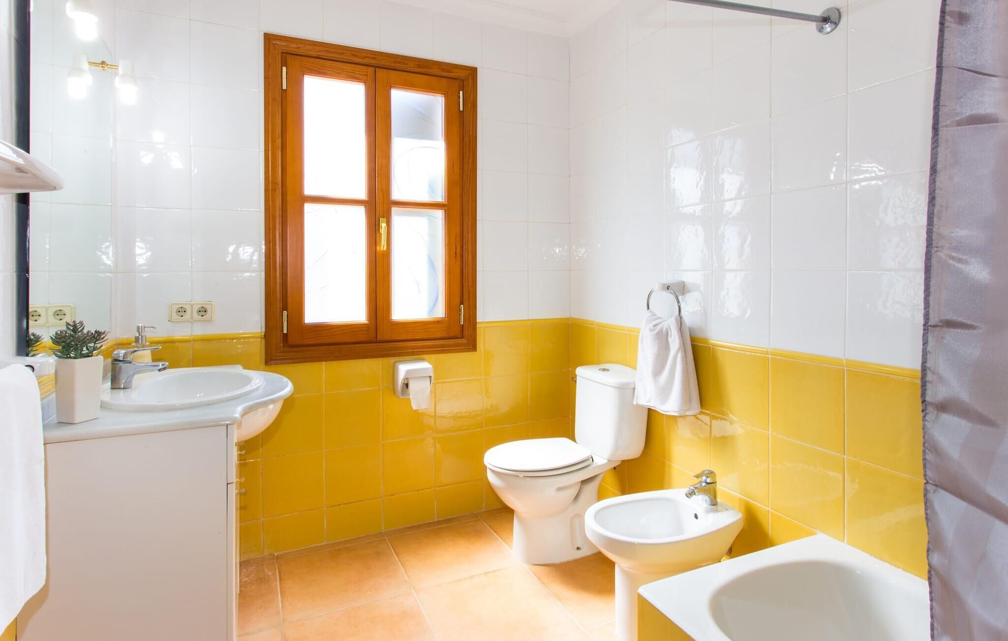 Een badkamer met een wand betegeld in geel en wit, een raam met houten kader, een wit wastafel, een toilet en een bidet.