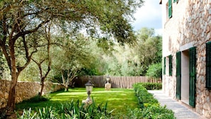 Un bâtiment en pierre avec des volets verts, un mur en pierre et un jardin bien entretenu comprenant une pelouse, une lanterne en pierre et diverses plantes.