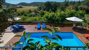 Ein Swimmingpool mit Liegestühlen und Sonnenschirmen, umgeben von Grünpflanzen und Blick auf Hügel.