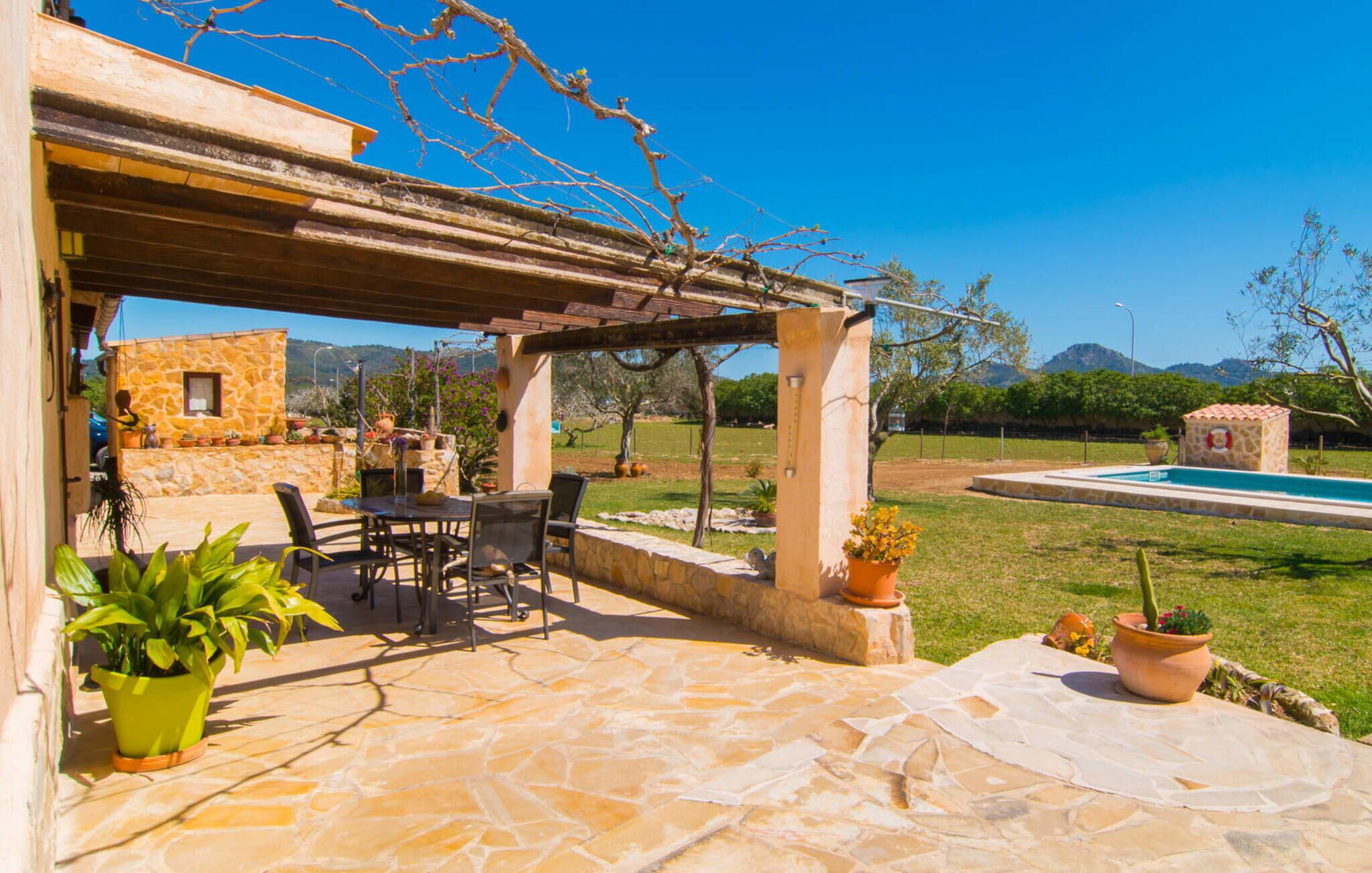 A stone patio with outdoor furniture, a pergola, a swimming pool, and a stone wall with potted plants.