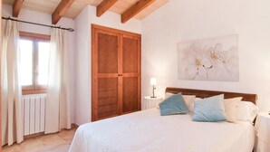 A bedroom with a wooden ceiling, a large bed with white bedding and blue pillows, a wooden wardrobe, a window with curtains, and a wall-mounted lamp. - Luxurious home in Son Servera (Son Servera)