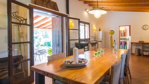 Una mesa de comedor de madera con sillas, un tazón de nueces y vasos de bebida verde. Se observa una cocina con un fregadero y un refrigerador.