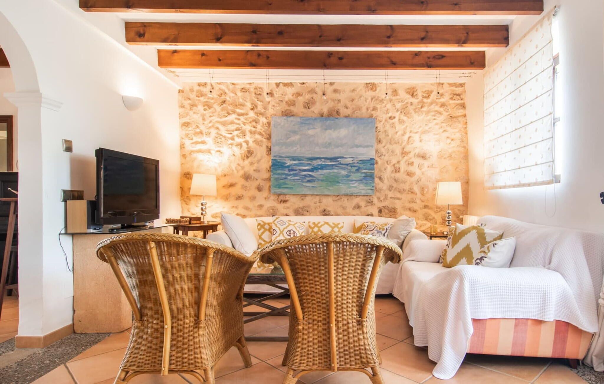 A cozy living room with a stone wall, a painting of the sea, wicker chairs, a sofa, and a TV.