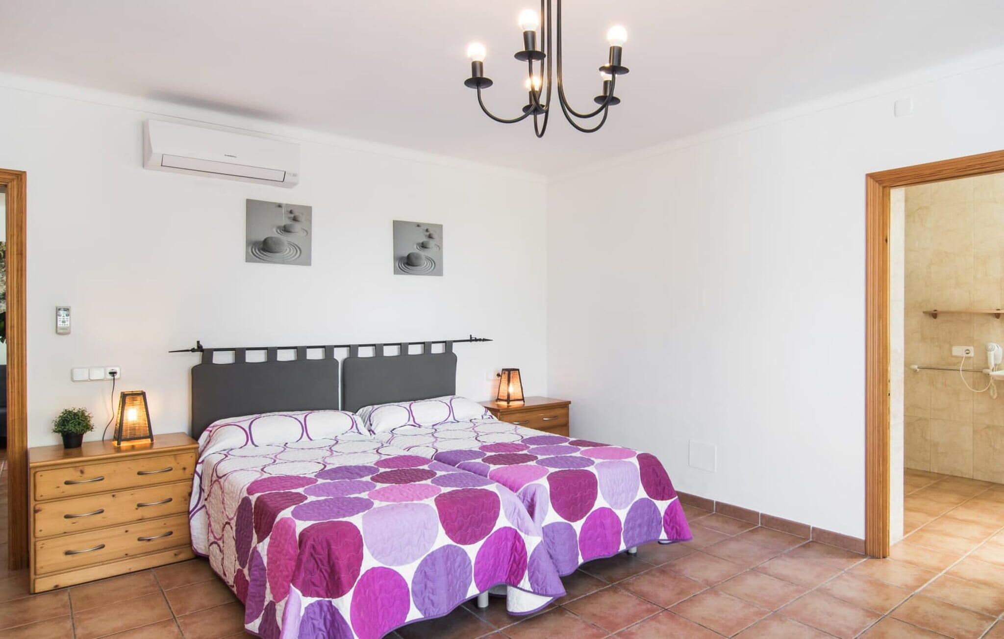 A bedroom with a bed, bedside tables, a chandelier, wall-mounted air conditioning, and artwork.