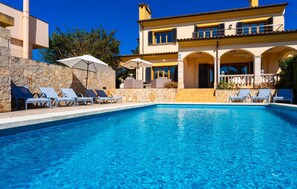 A swimming pool with lounge chairs and umbrellas, a stone wall, and a yellow building with balconies. - Cozy home in Cala Millor with private swimming pool, can be inside or outside (Cala Millor)