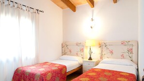 A room with two single beds, each with floral-patterned headboards and red bedspreads. There is a nightstand with a lamp between the beds, and a window with sheer curtains on the left. - Lovely home in Colonia de Sant Jordi (Colonia de Sant Jordi)