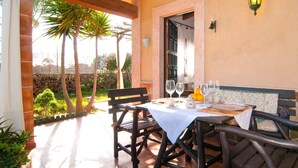 An outdoor dining area with a table set for two, surrounded by wooden chairs, a stone wall, and palm trees. - Lovely home in Colonia de Sant Jordi (Colonia de Sant Jordi)