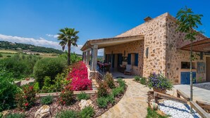 Una casa de piedra con un porche cubierto, rodeada por un jardín con diversas plantas y vistas al campo.