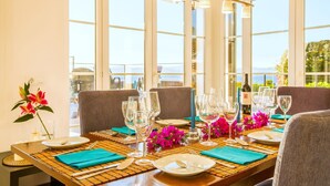 Mesa de comedor preparada para seis personas, con cristalería transparente, platos y cubiertos, un centro de mesa con velas, y vista al mar a través de grandes ventanales.