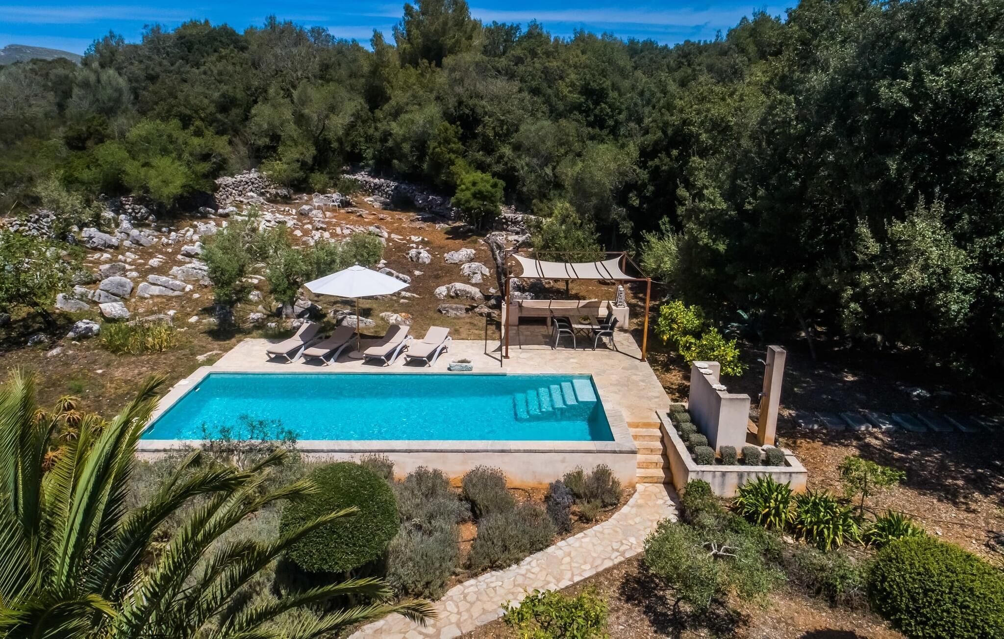Une piscine rectangulaire, agrémentée de chaises longues et d’un espace salon couvert, le tout entouré d’un jardin aménagé et d’arbres.