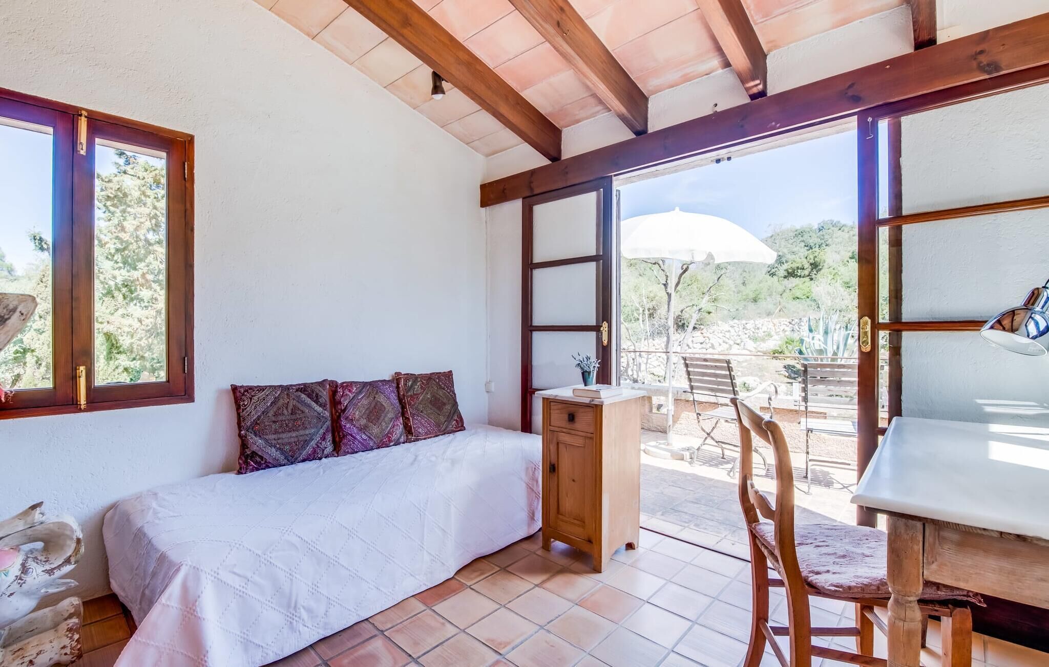 A room with a bed, a wooden cabinet, a table, and a chair. There is a window and a door leading to an outdoor area with a table and chairs.
