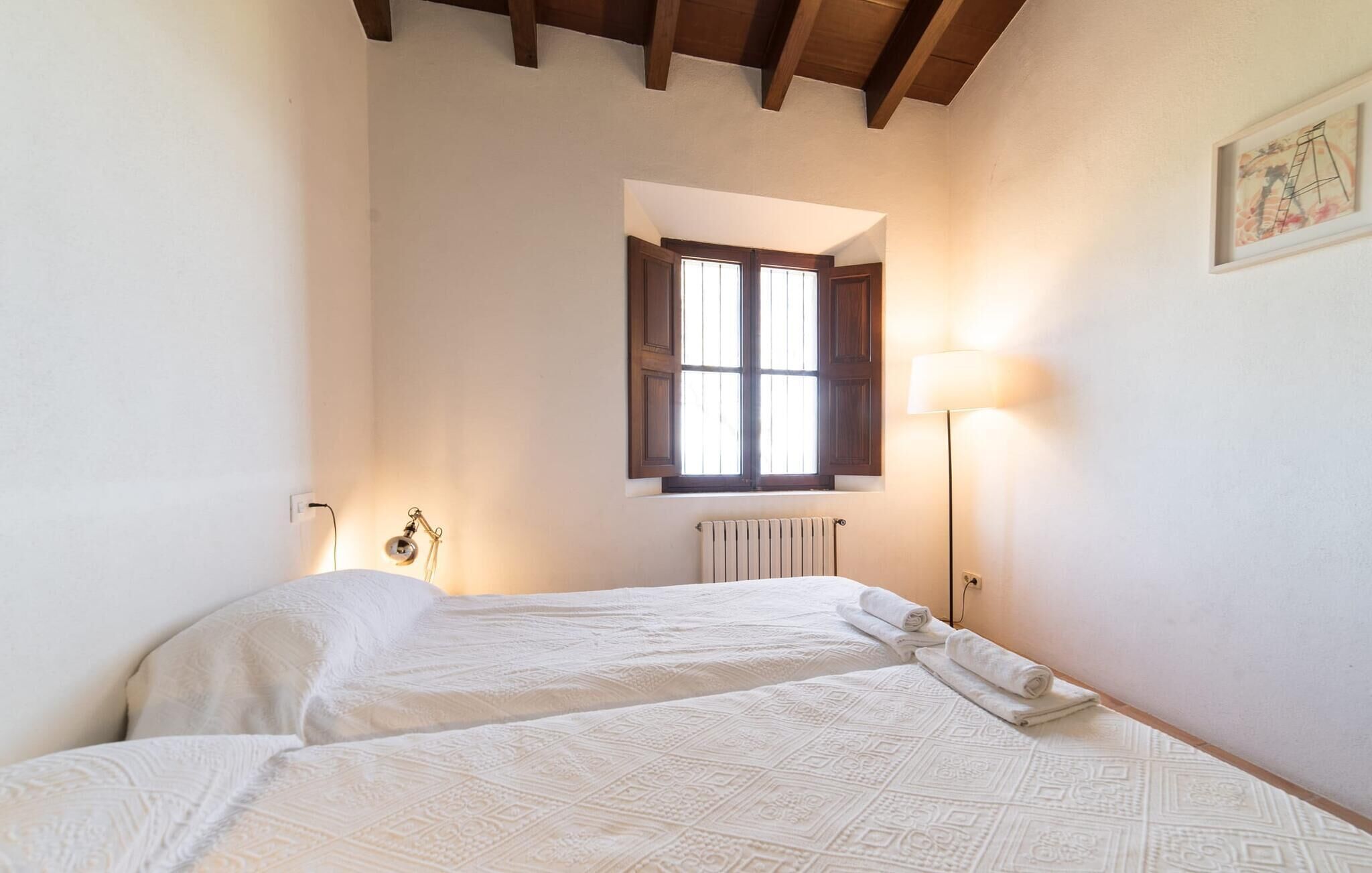 A bedroom with two beds, a window with wooden shutters, a radiator, and a wall-mounted lamp.