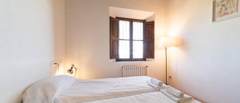 A bedroom with two beds, a window with wooden shutters, a radiator, and a wall-mounted lamp.