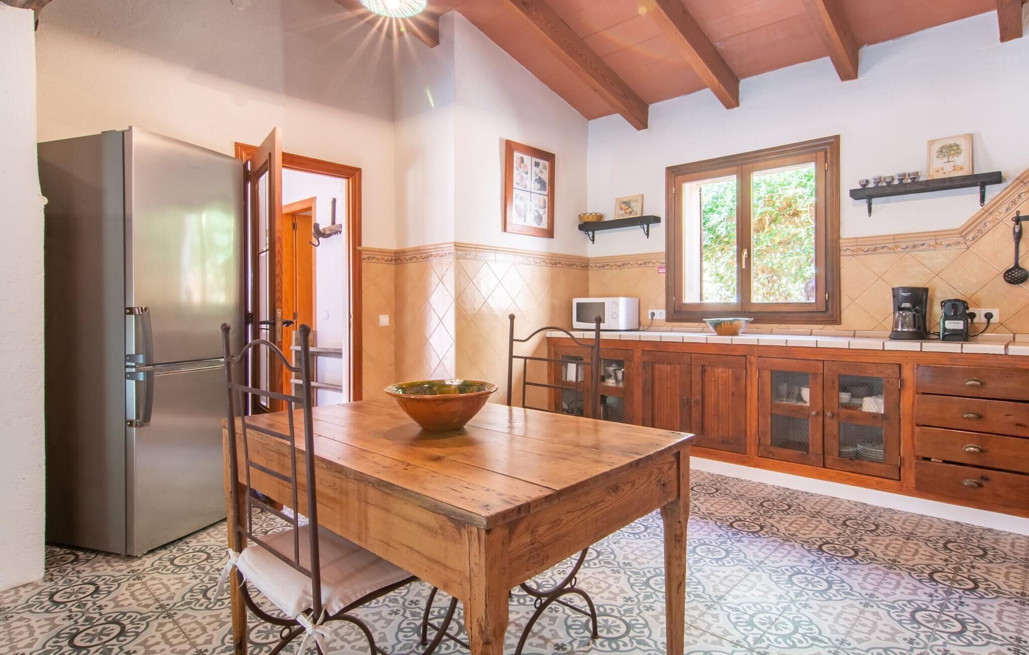 A kitchen with a wooden table, chairs, a refrigerator, cabinets, a microwave, and a window with a view of greenery.