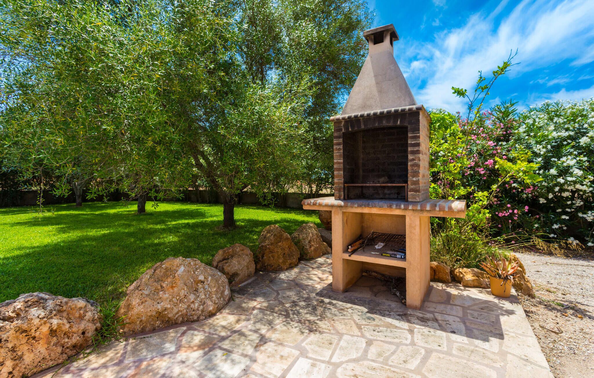 An outdoor brick barbecue grill with a chimney, set on a stone patio surrounded by rocks and greenery.