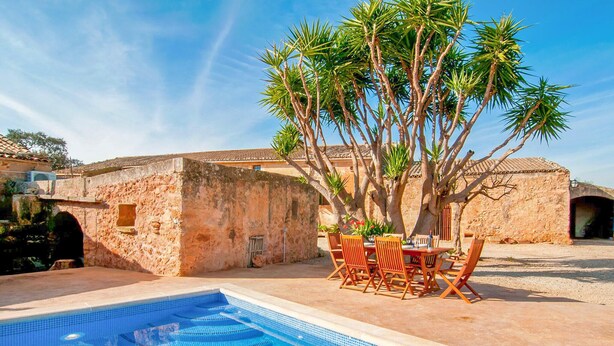 Una piscina con borde de azulejos, una mesa de madera con sillas, y un árbol con follaje verde.
