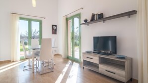 Ein modernes Wohnzimmer mit einem Glastisch, weißen Stühlen, einem Fernseher auf einem Holzboden und einer grün gerahmten Tür, die nach draußen führt.