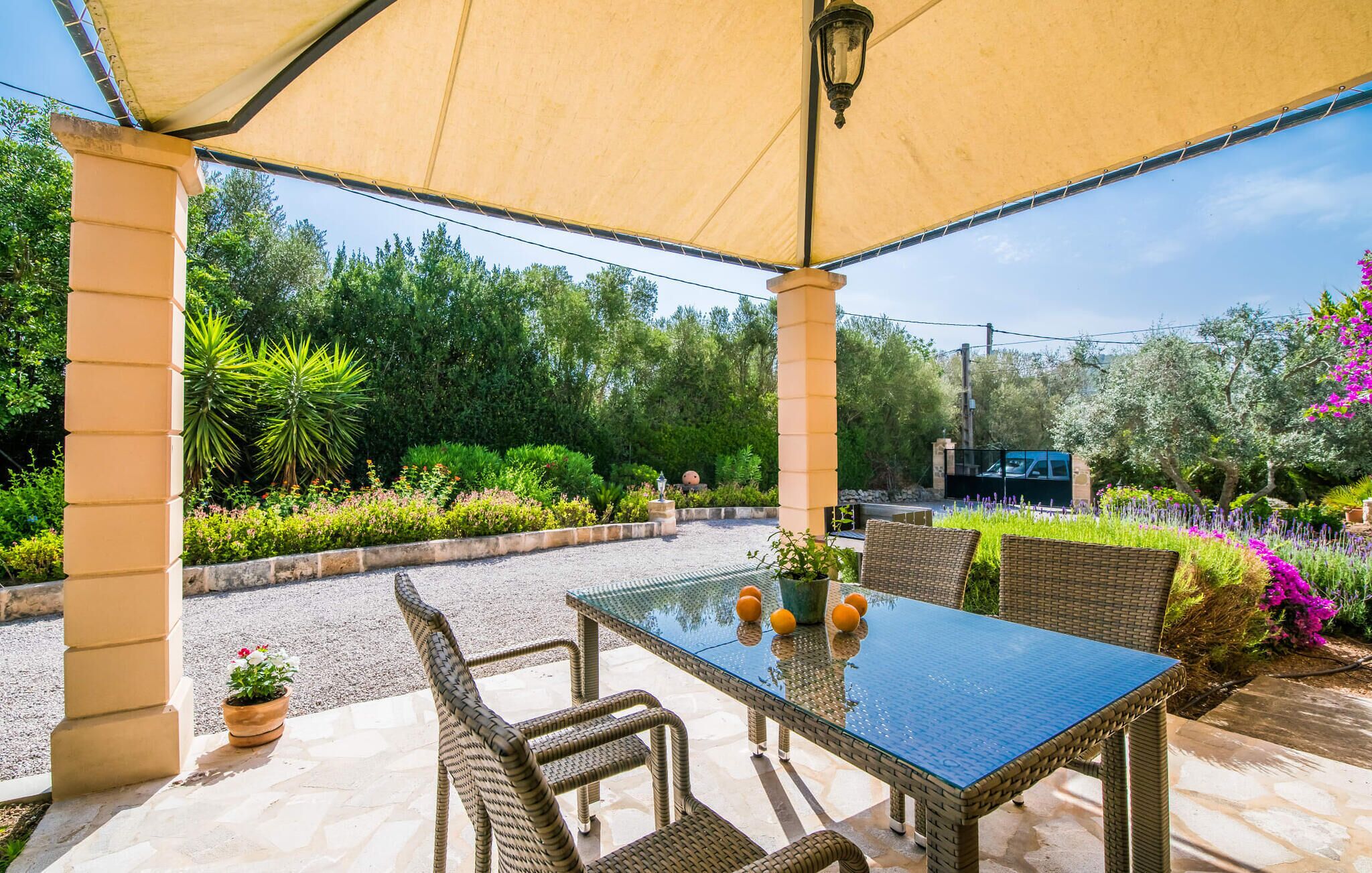 Un patio al aire libre con una mesa de vidrio y sillas de mimbre, rodeado de abundante follaje y una lámpara decorativa.