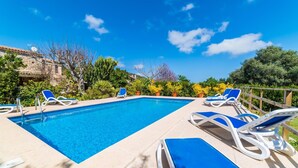 Ein rechteckiger Swimmingpool mit klarem, blauem Wasser, umgeben von einer Betonplattform. Mehrere weiße Liegestühle mit blauen Kissen sind um den Pool angeordnet. Die Plattform wird von einem Holzzaun begrenzt, im Hintergrund sind verschiedene Kübelpflanzen und Bäume zu sehen.
