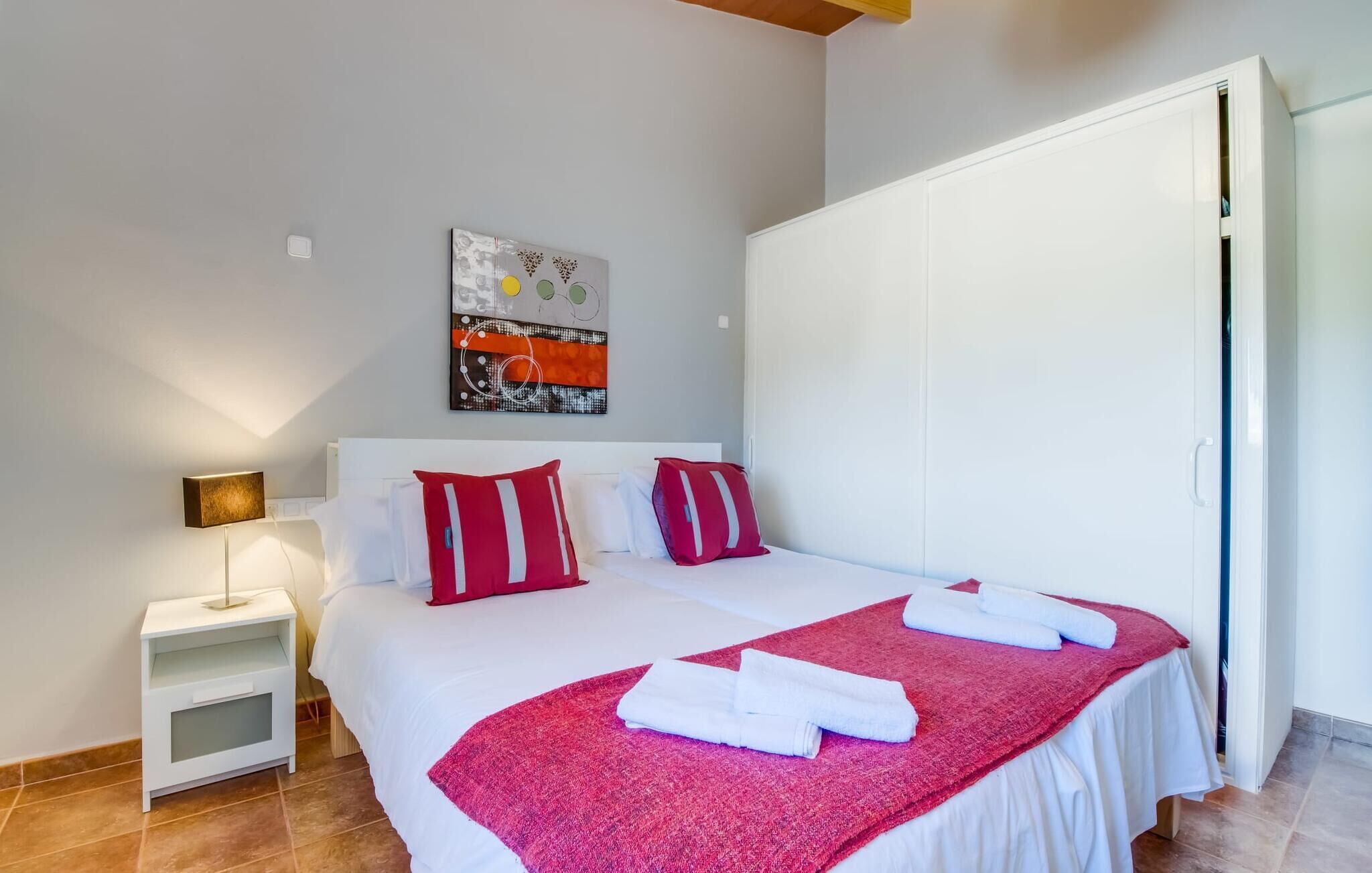 Un lit bien fait, avec du linge de lit blanc et des coussins rouges, une petite table de chevet avec une lampe, un plaid rouge et un tableau accroché au mur.