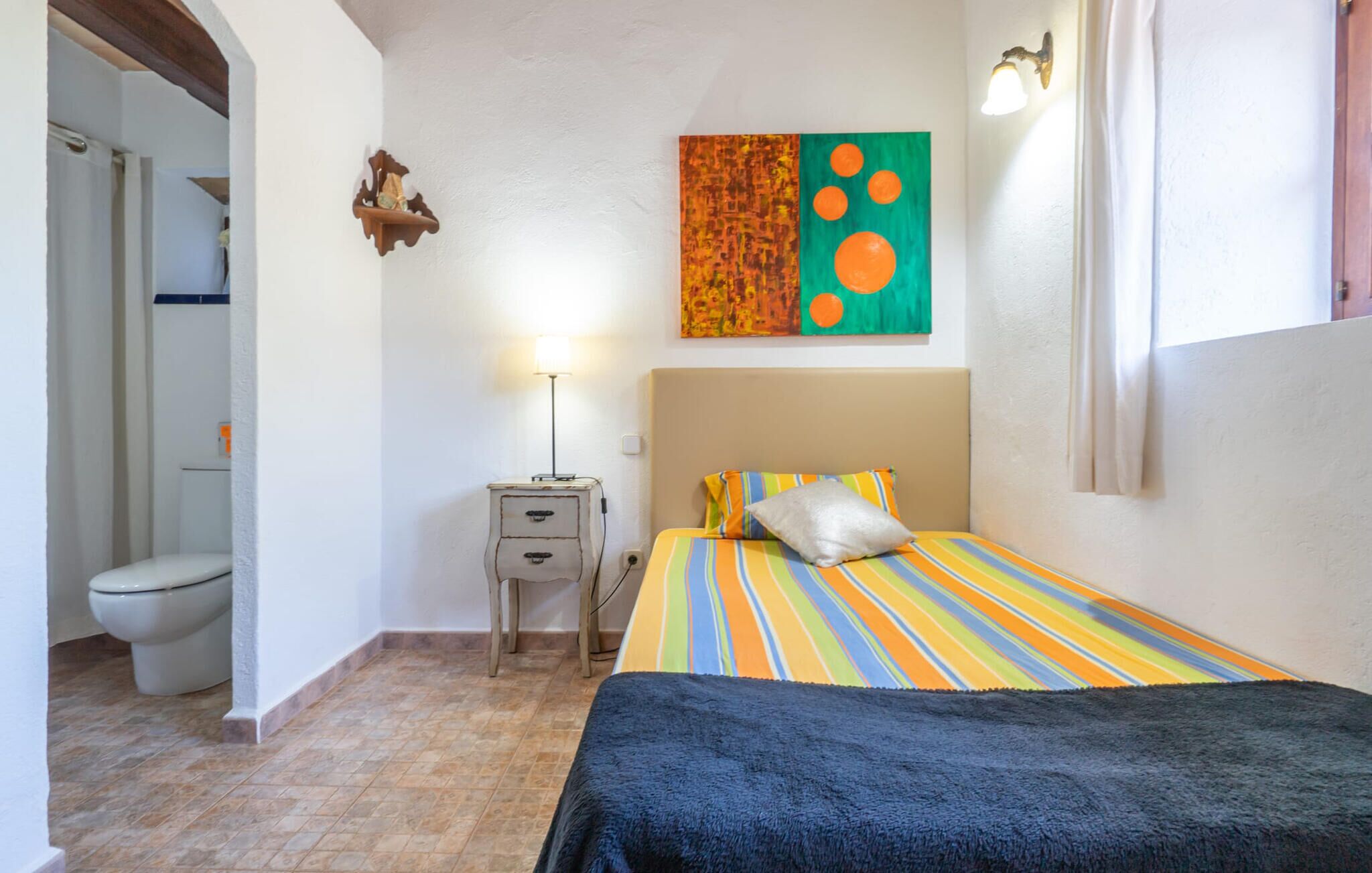 A bedroom with a bed, a bedside table, a lamp, a painting, and a bathroom visible through an archway.
