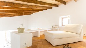 A bright, white room with a white sofa, a small table, and a bench. There is a window letting in natural light, and a pair of sandals on the floor. - Nice home in Sant Llorenc with private swimming pool, can be inside or outside (Sant Llorenc)