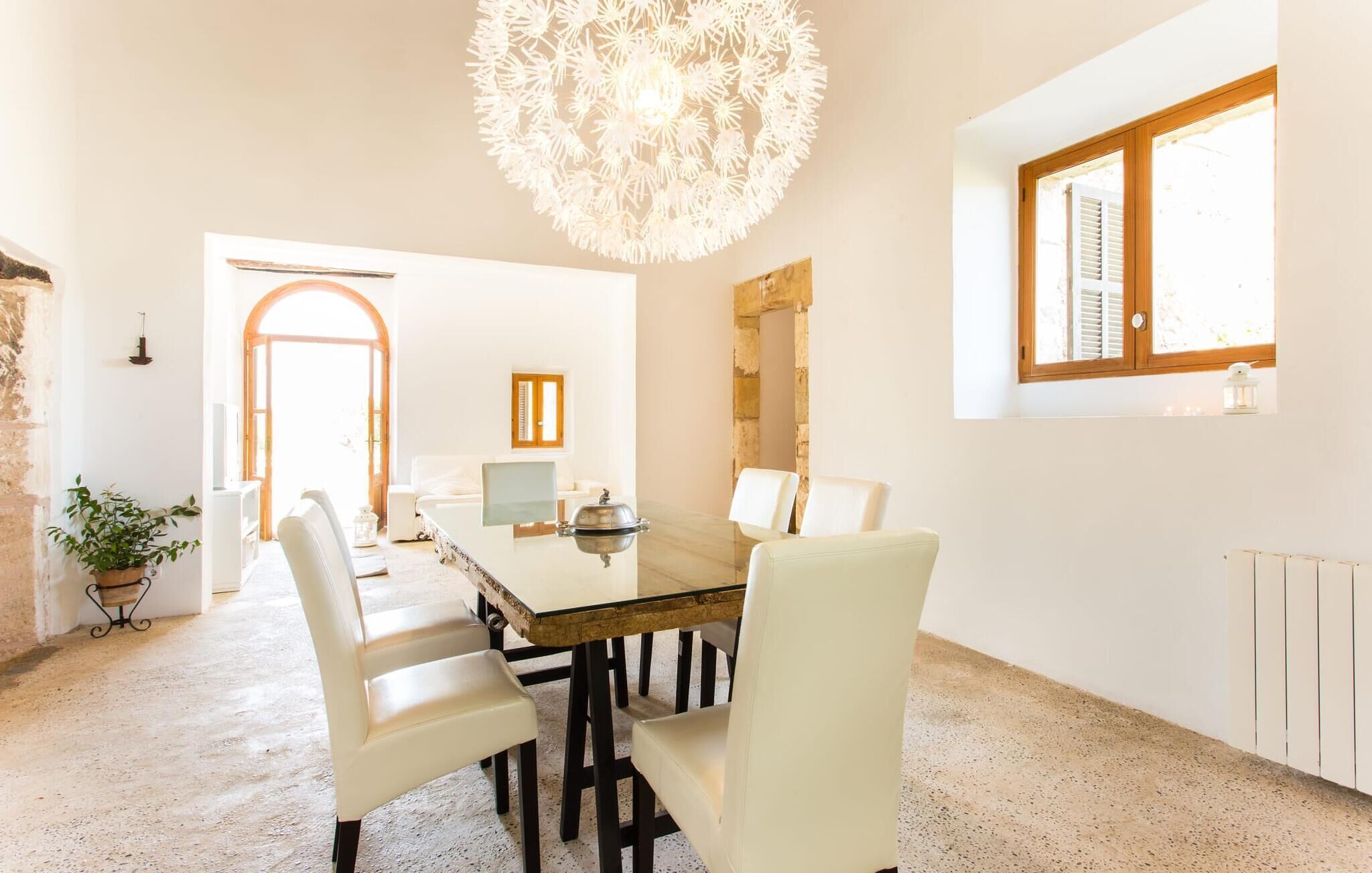 A spacious dining area with a large table, white chairs, a chandelier, and a view of the outdoors through a window.