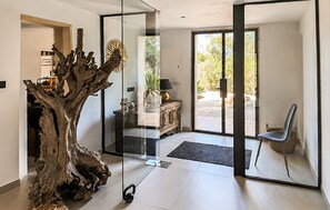 A modern interior with a glass door leading to an outdoor area, a wooden tree sculpture, and a seating area with a chair and a small table. - Luxurious home in Portocolom with sauna (Portocolom)