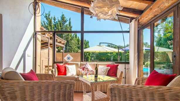 Un patio iluminado por el sol con muebles de mimbre, una mesa de vidrio y vistas a una piscina y árboles.