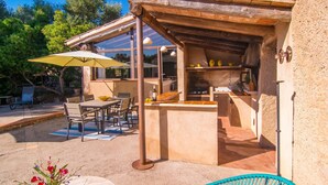 Un patio exterior con mesa y sillas, un paraguas amarillo y una zona de barbacoa con parrilla y estantes.