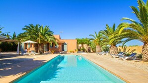 Piscina com espreguiçadeiras e palmeiras, cercada por um edifício com telhado telhado.