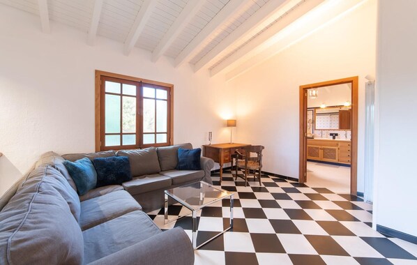 Uma sala de estar com piso xadrez, um sofá, uma mesa de centro de vidro e uma escrivaninha com uma cadeira.