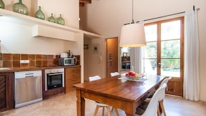 Eine moderne Küche mit einem Holztisch und Stühlen, einer hängenden Leuchte und Blick ins Freie durch ein großes Fenster.