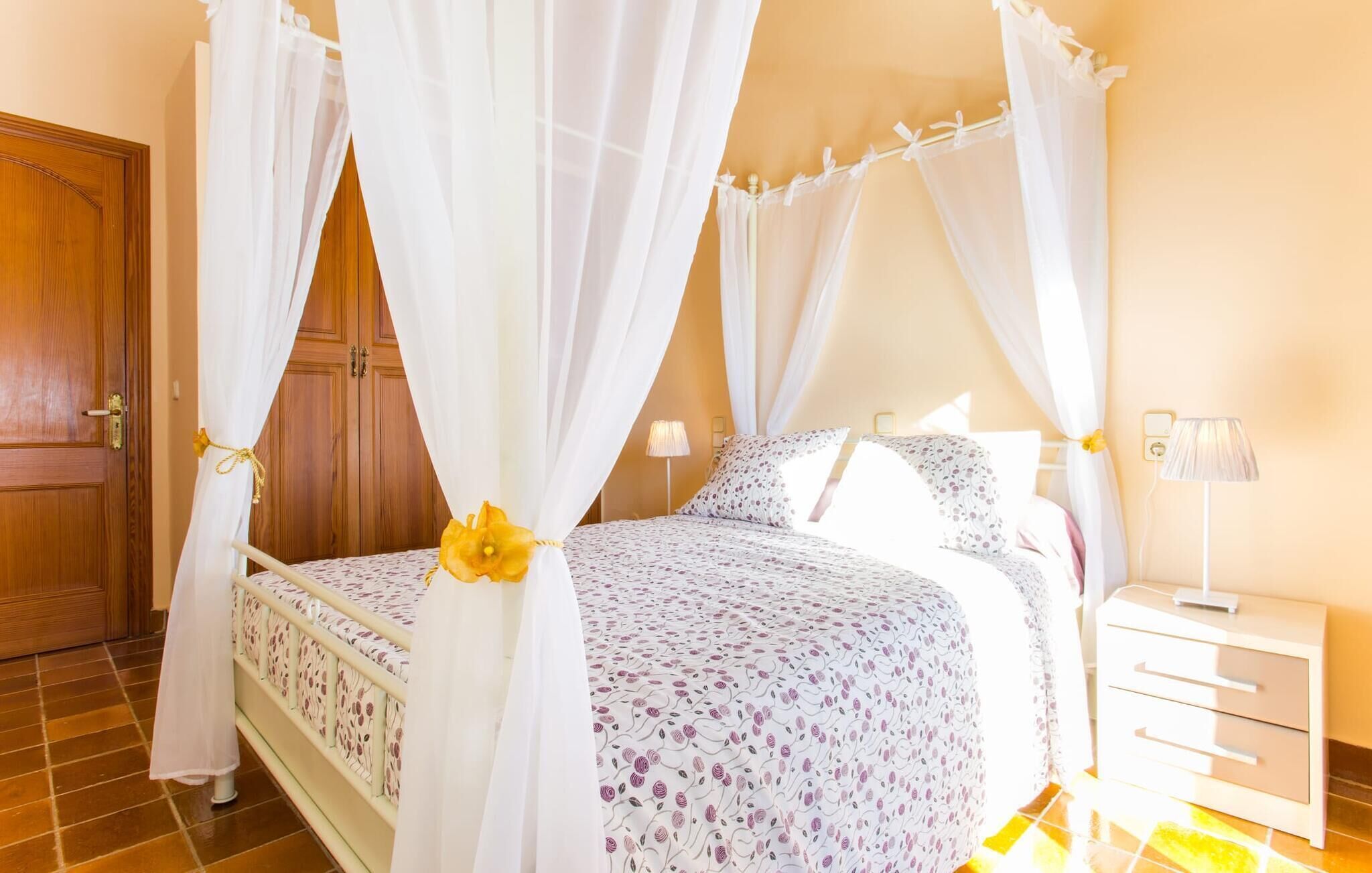 A bedroom with a four-poster bed, white curtains, a wooden door, a bedside table with a lamp, and a tiled floor.