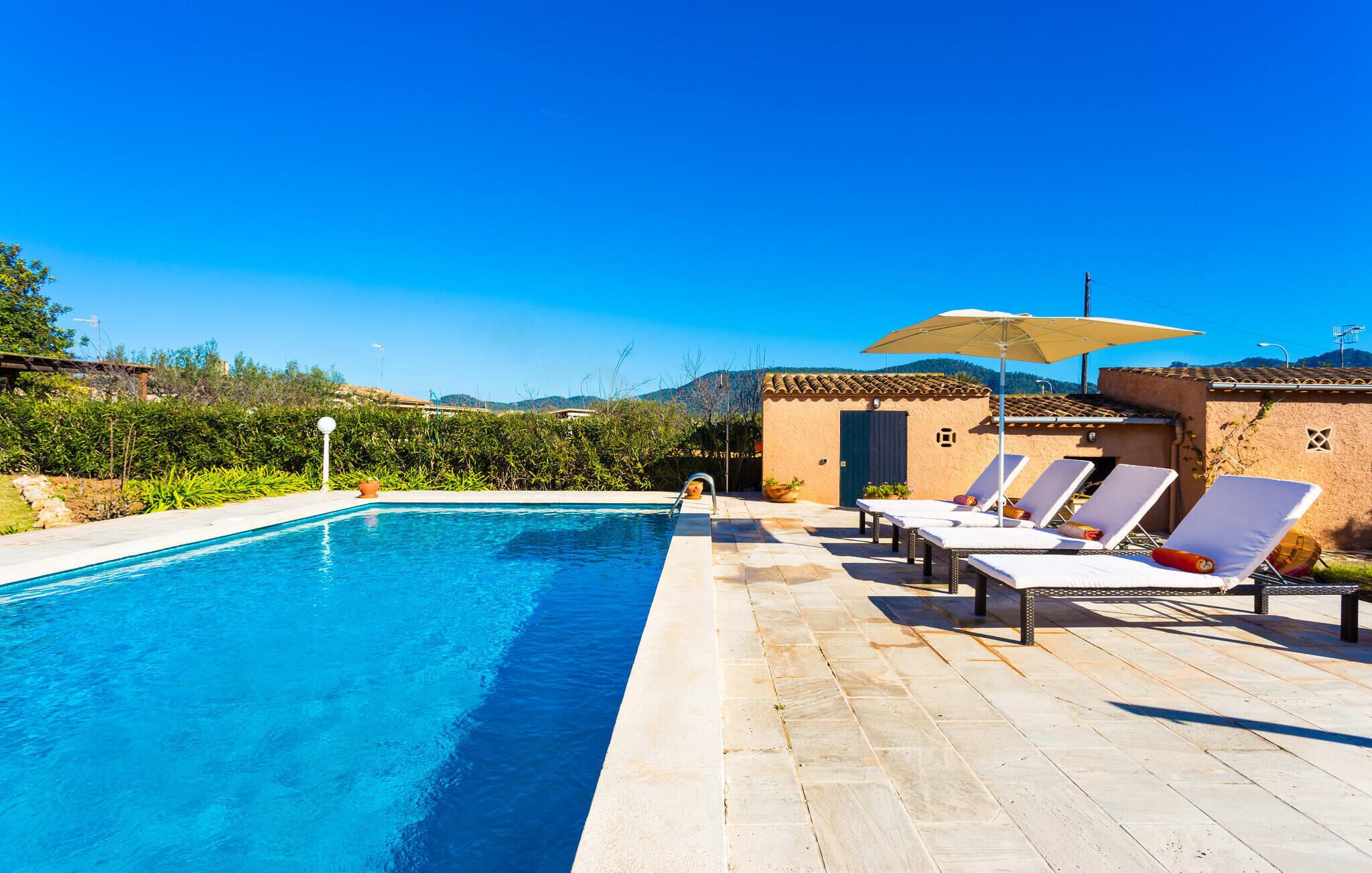 Une piscine entourée de chaises longues et de parasols, elle-même bordée d’un jardin aménagé et d’un bâtiment doté d’un toit en tuiles.