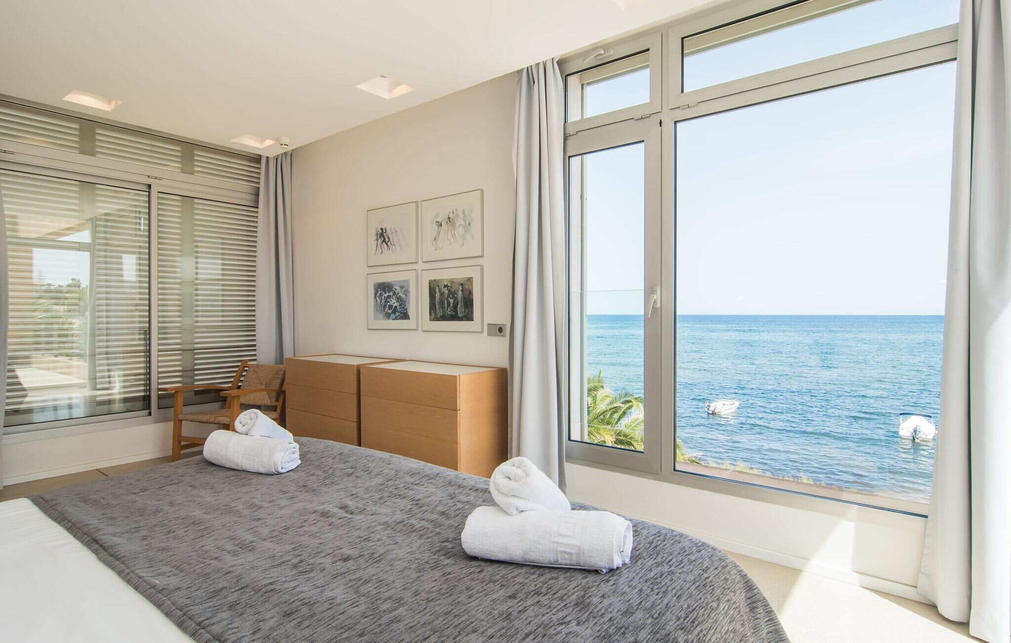 A modern bedroom with a large window overlooking the sea, a bed with two white towels, and a wooden headboard.