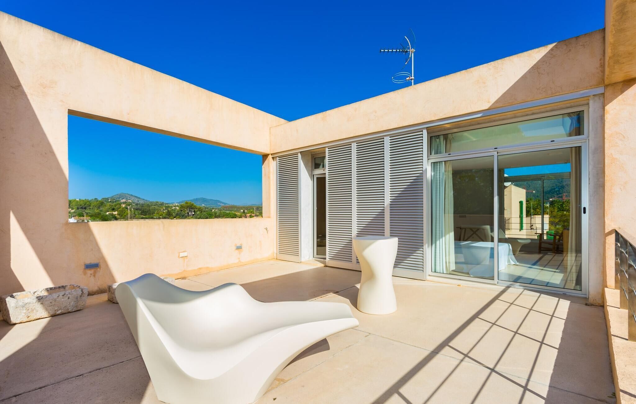 Eine moderne Terrassenanlage mit einem weißen, wellenförmigen Stuhl und einem kleinen Tisch, einer Glasschiebetür, die zu einem überdachten Bereich führt, und freiem Blick auf eine Berglandschaft.