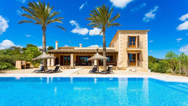 Un edificio de piedra de dos pisos con piscina, palmeras y mobiliario exterior.