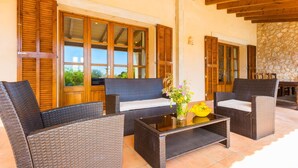 Un patio con mobiliario de mimbre, una mesa de centro de vidrio y contraventanas de madera en las ventanas.