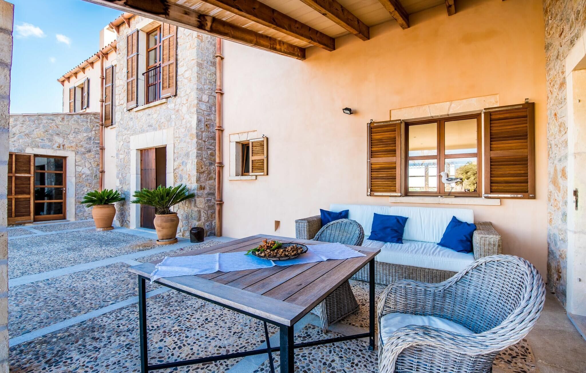 A cozy outdoor seating area with a table and wicker furniture, stone walls, and wooden shutters.