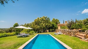 Une piscine rectangulaire bordée de pierres, entourée d’une pelouse bien entretenue et d’arbres. Des chaises longues et des parasols sont disposés sous les arbres.