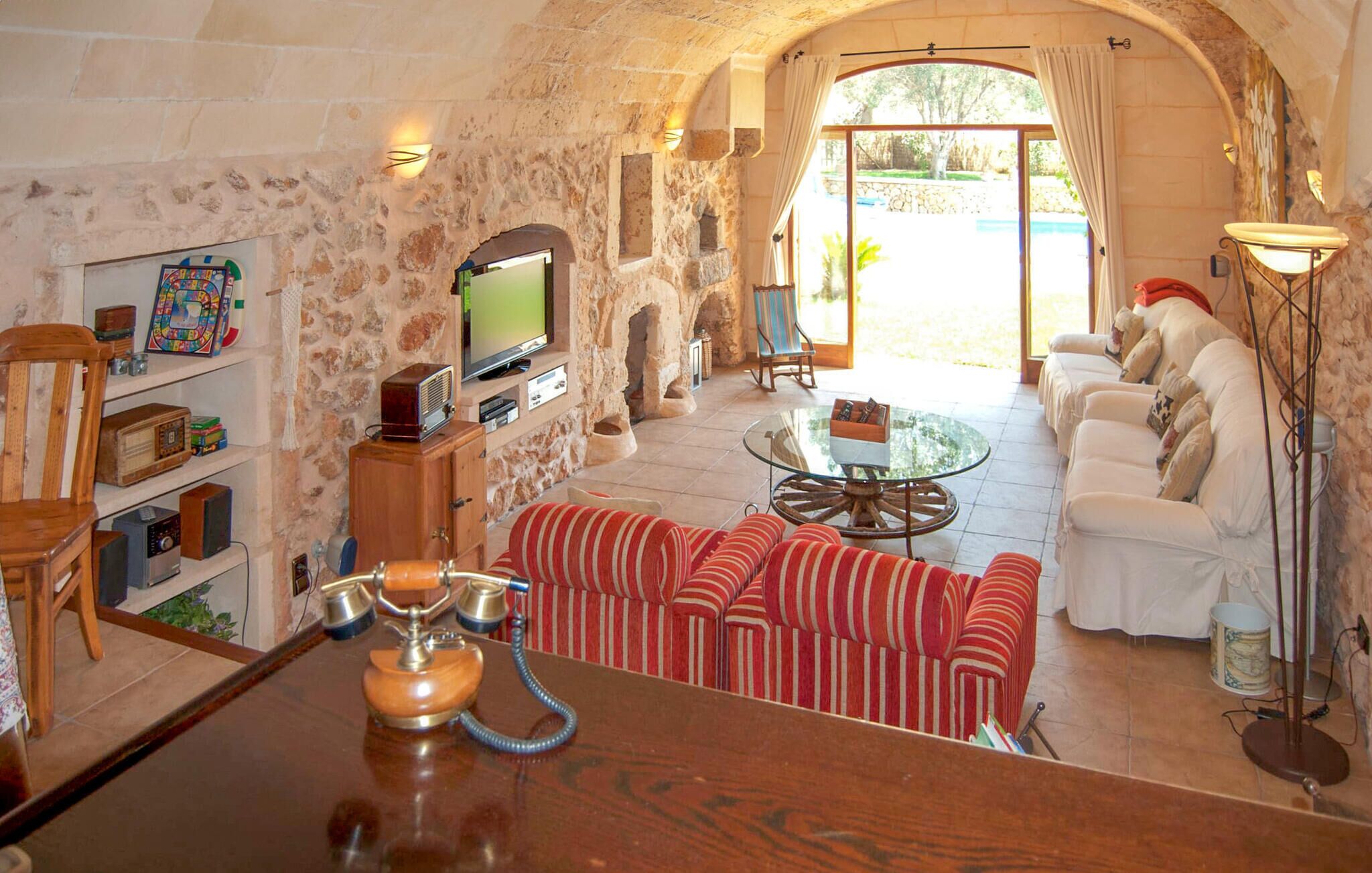 A stone-walled room with a television, a wooden cabinet with vintage radios, striped red and white sofas, and a glass coffee table.