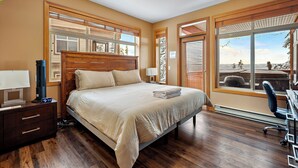 Ein Schlafzimmer mit einem großen Bett, einem hölzernen Kopfteil, Nachttischen, einem Schreibtisch mit Stuhl und einem Fenster mit Blick auf Bäume und ein Gebäude.