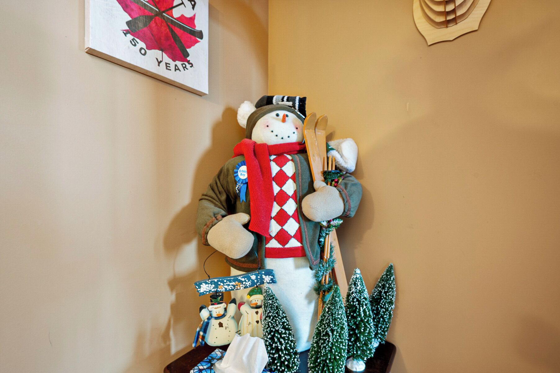 Un adorno de muñeco de nieve con bufanda y gorro, rodeado de pequeños abetos navideños y otras decoraciones.